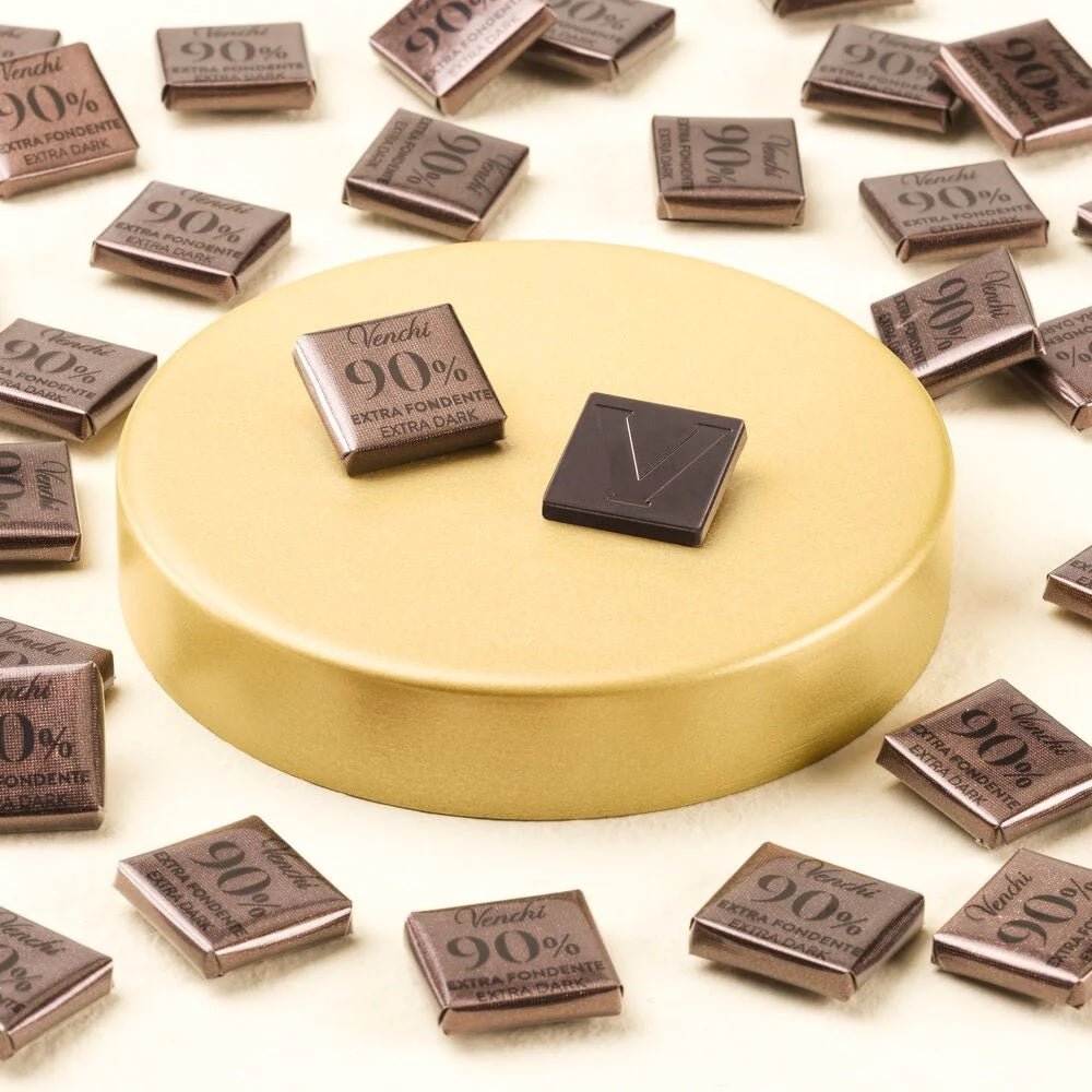 A round gold pedestal surrounded by scattered Venchi 90% chocolate squares, with two chocolate pieces placed on top: one wrapped in brown paper and one unwrapped, showing a dark brown chocolate surface with a V logo.