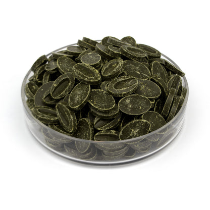 Dark olive green oval chocolate feves piled in a clear glass bowl on a white background.
